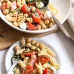 a small plate with chickpea pasta salad along with a serving bowl full of the salad, with tomatoes, bocconcini, salami and basil