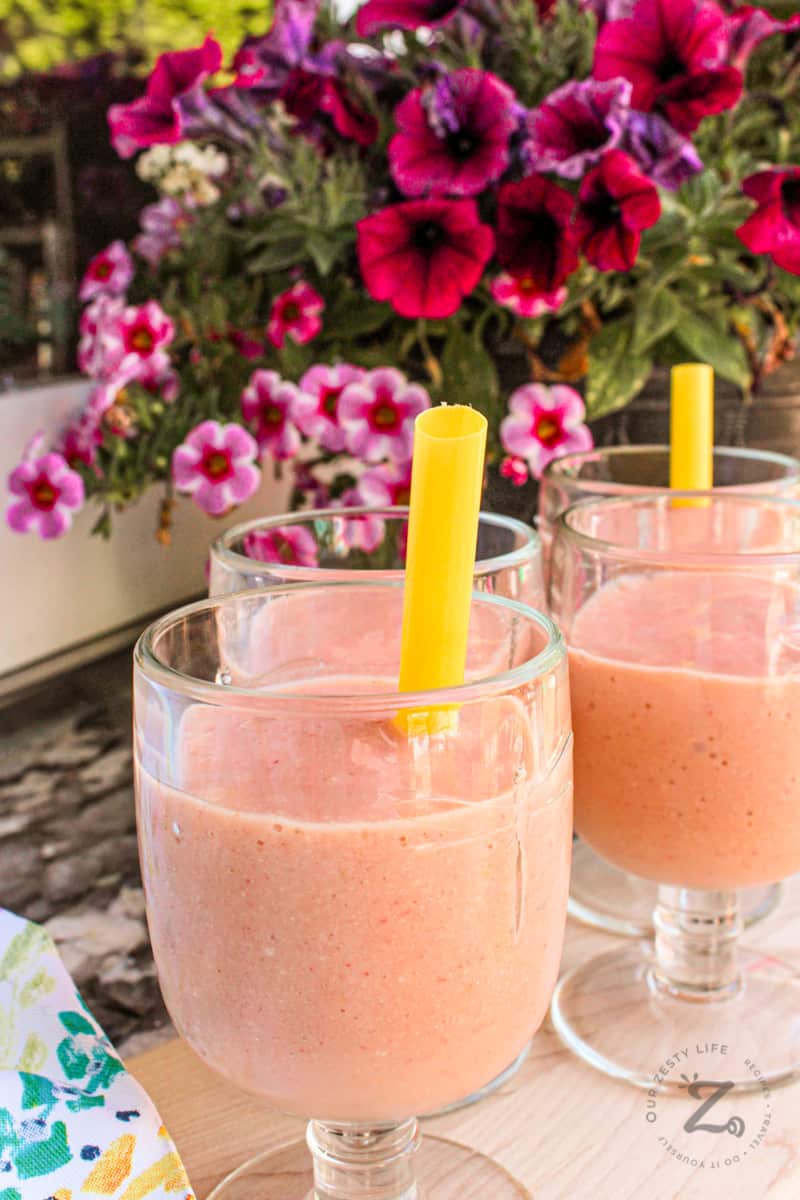 Four glasses of strawberry mango smoothies on a serving tray with yellow straws and flowers in the background