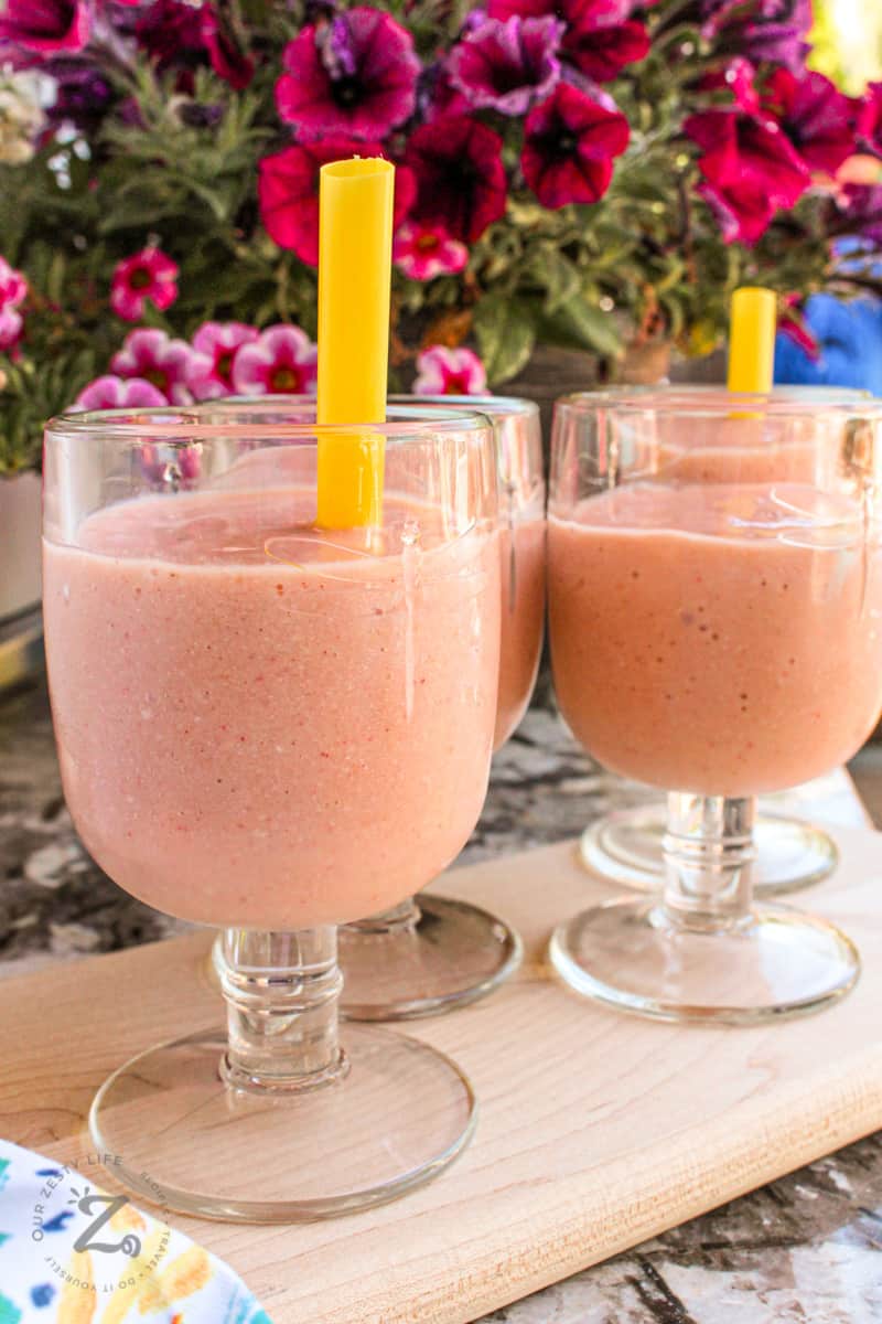 A close up of four strawberry mango smoothies on a tray with straws in them and flowers in the background