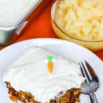 piece of carrot cake on a white plate with a fork with a bowl of pineapple on the side