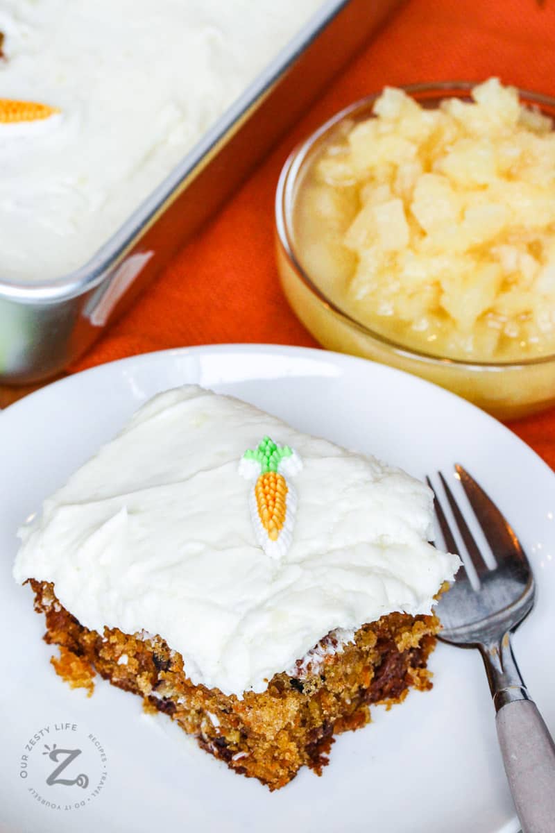 Piece of carrot cake on a plate with a fork, with a bowl of pineapple on the side 