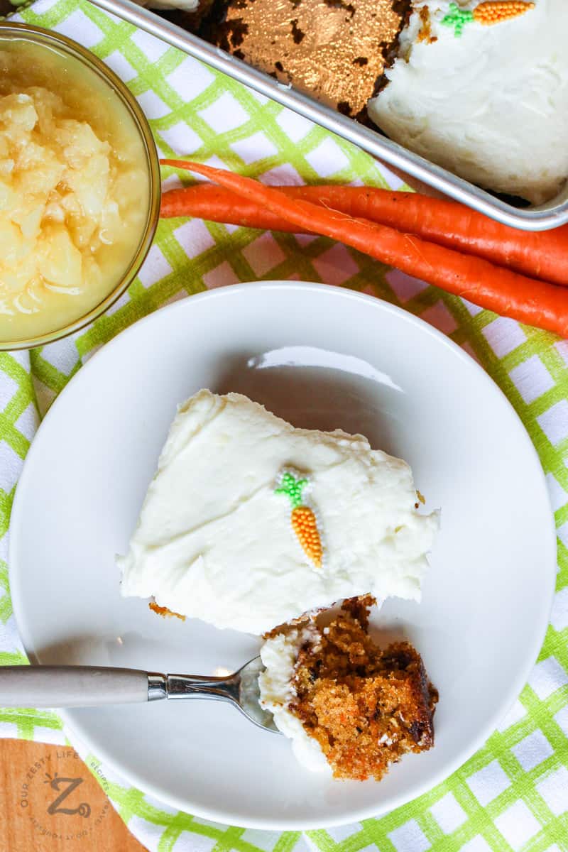 Piece of carrot cake on a plate with a bite on a fork, with carrots, pineapple and the rest of the cake on the side 
