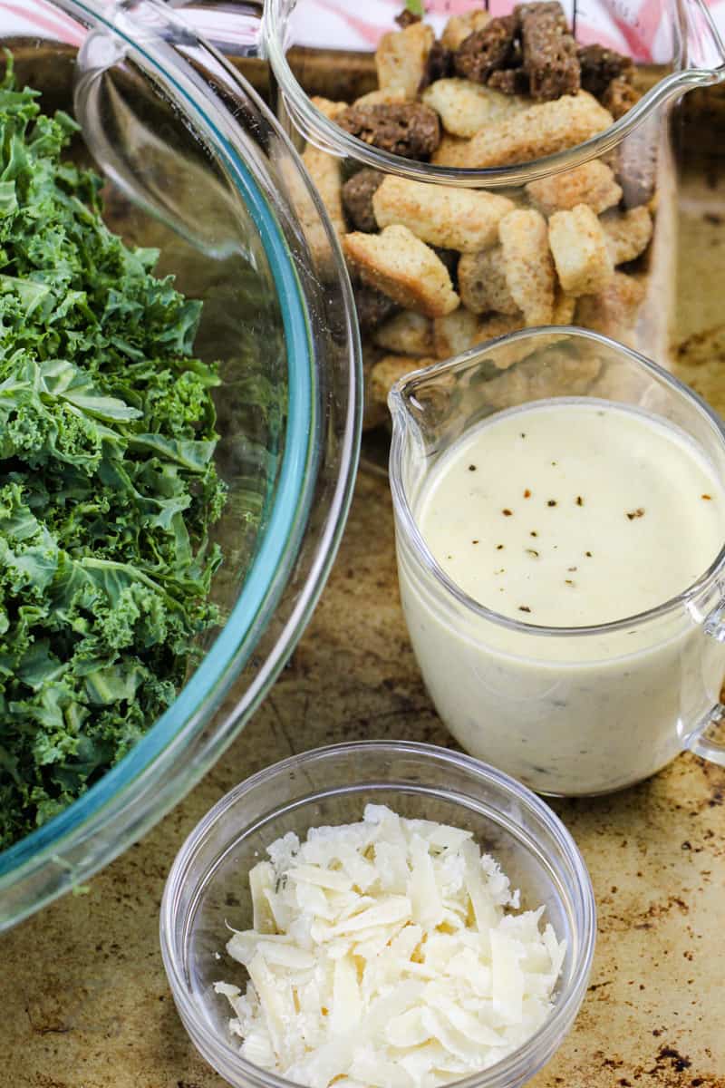 kale croutons cheese and dressing in bowls to make Kale Caesar Salad