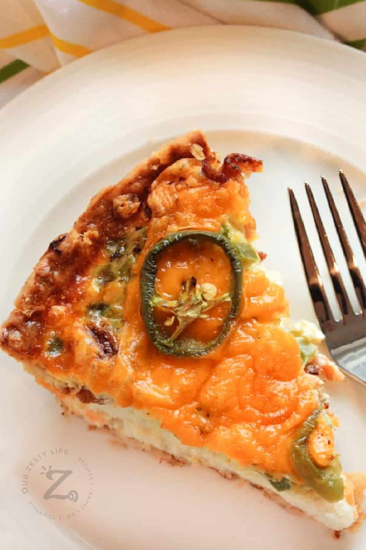 close up of zesty bacon and cheese quiche, with a jalapeno slice on top, on a white plate with a fork on the side.