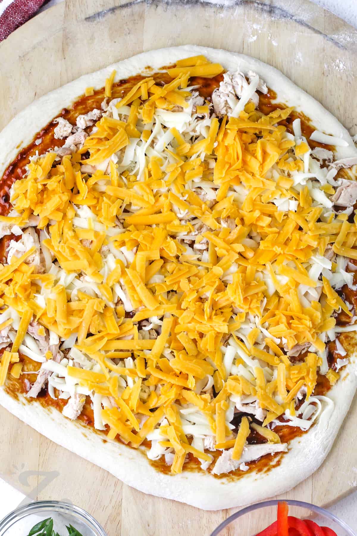 a cheese layer added to a whole unbaked BBQ chicken pizza on a wooden board