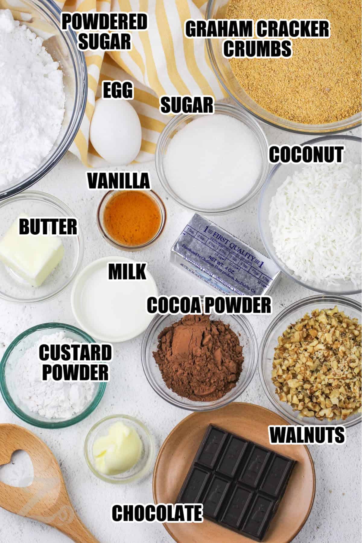 ingredients to make Nanaimo Bars including powdered sugar, graham cracker crumbs, egg, sugar, coconut, vanilla, butter, milk, cocoa powder, custard powder, walnuts, and chocolate