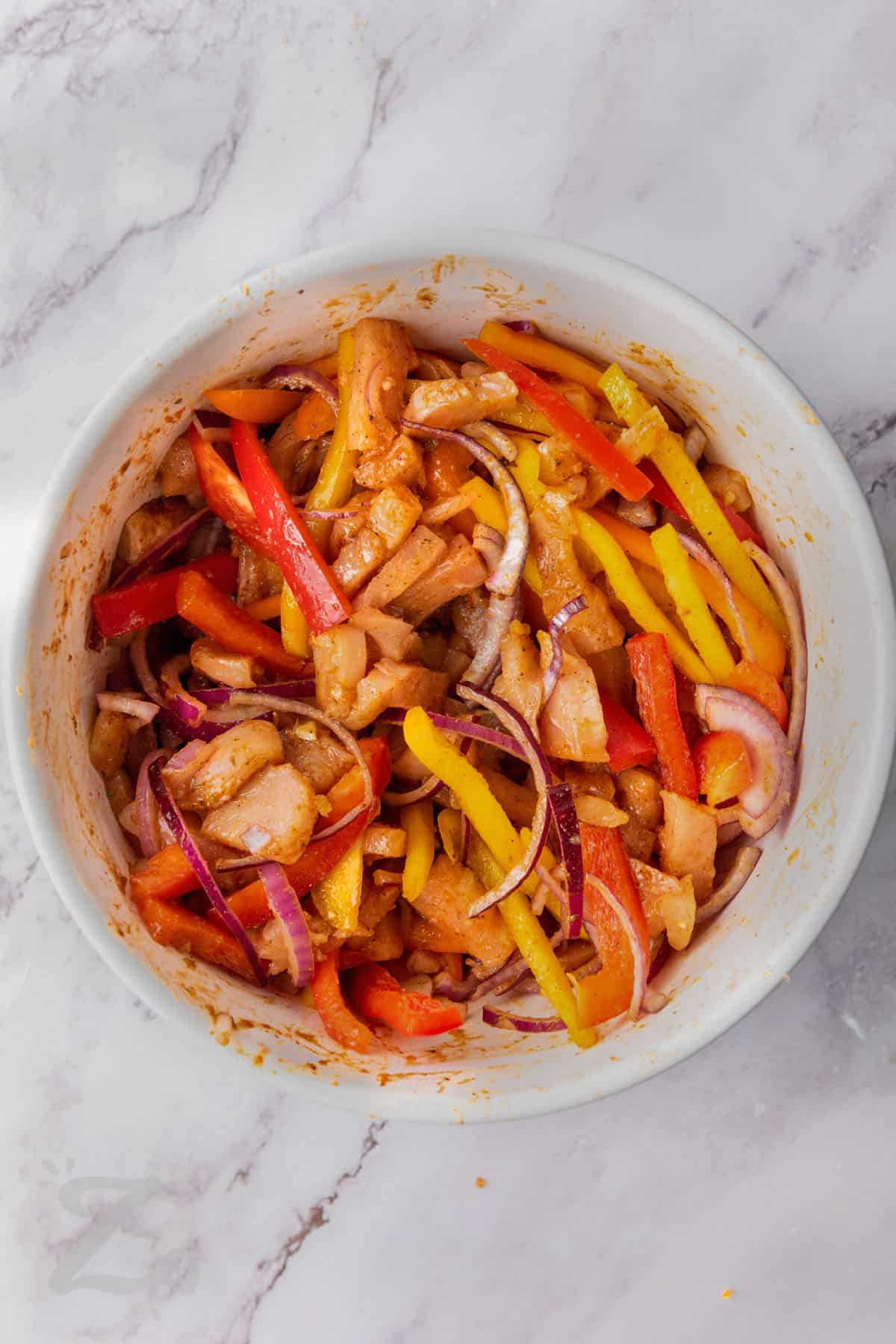 chicken breasts , red onions, and bell peppers tossed in seasoning in a bowl.