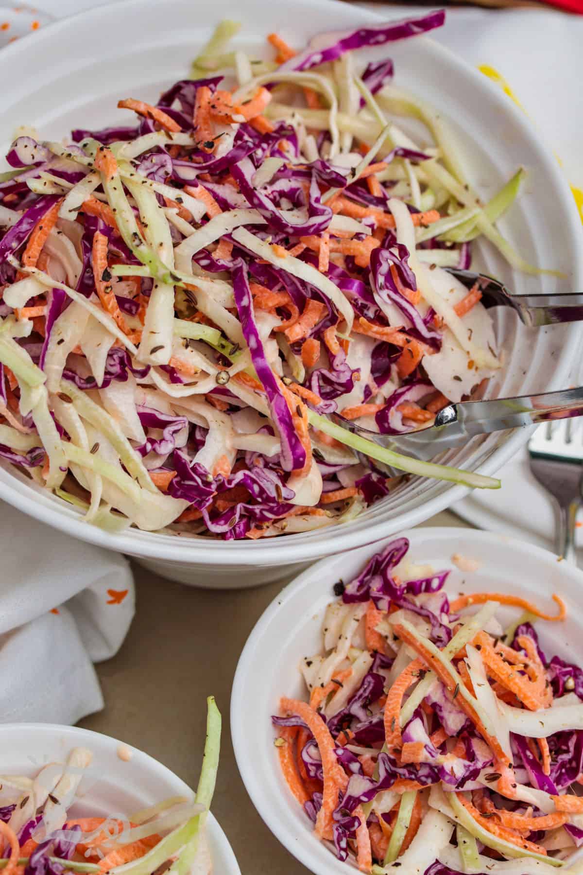 a large bowl of creamy coleslaw with silver tongs in it, with two serving bowls of coleslaw in front