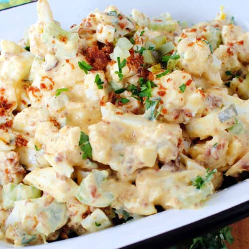 cauliflower potato salad in a white dish, garnished with chopped pickles, bacon bits and chopped parsley