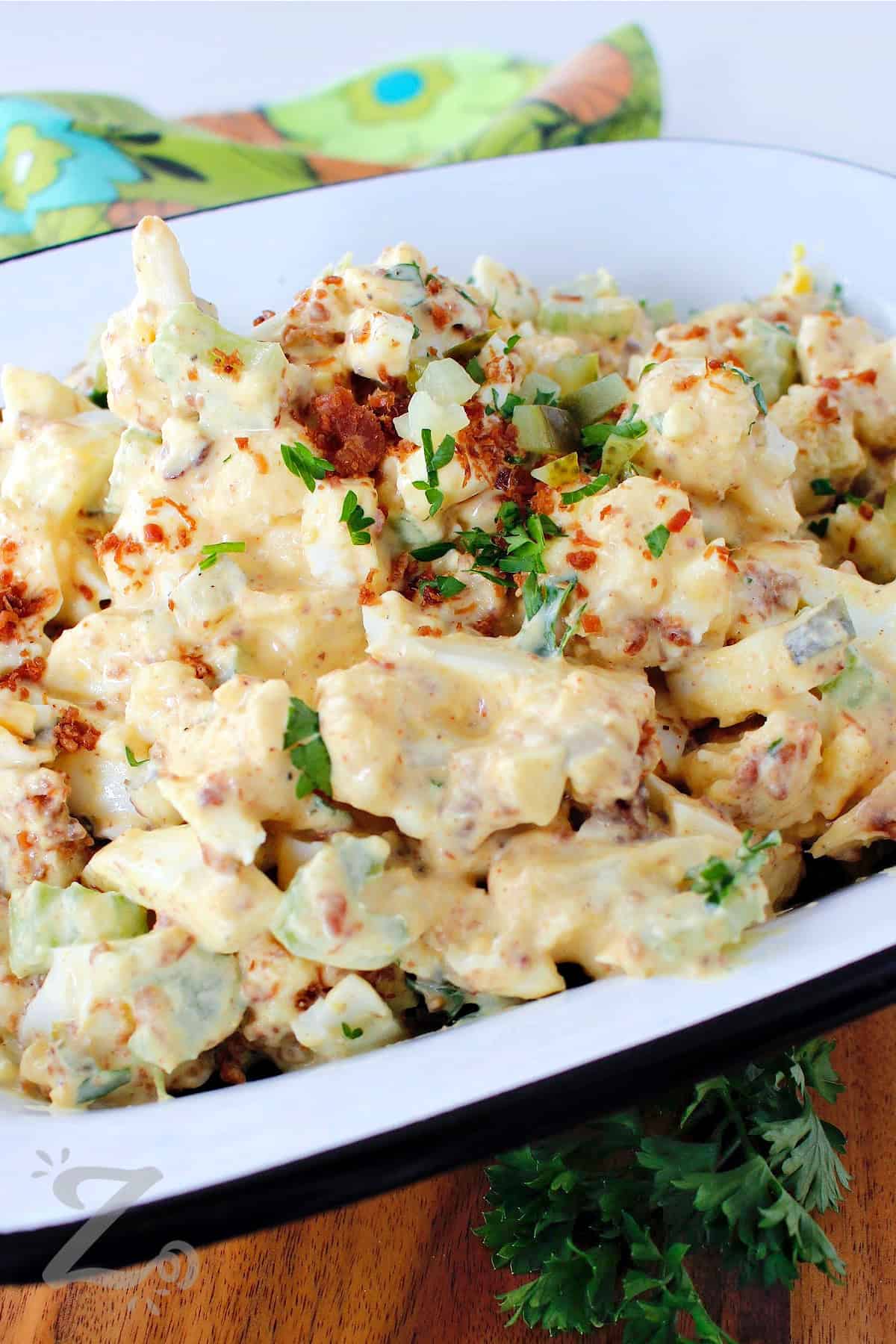 cauliflower potato salad in a white dish, garnished with chopped pickles, bacon bits and chopped parsley