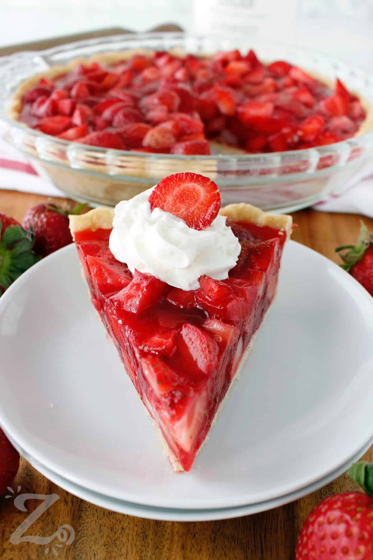 Fresh strawberry pie served with whipped cream and a sliced strawberry on top, on a small white plate, and the remaining pie in the background