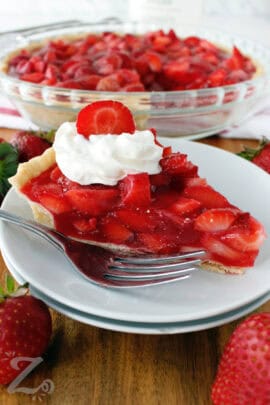 Fresh strawberry pie with whipped cream and a sliced strawberry on top, served on a small white plate with a fork, and the remaining pie in the background