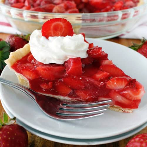 Fresh strawberry pie with whipped cream and a sliced strawberry on top, served on a small white plate with a fork, and the remaining pie in the background