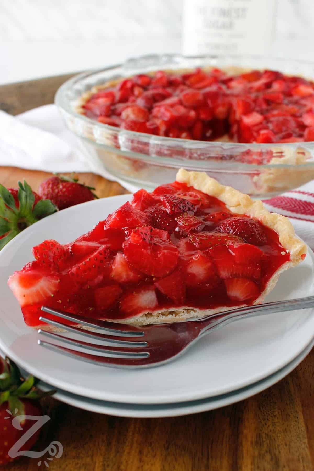 Fresh strawberry pie served on a small white plate with a fork, and the remaining pie in the background