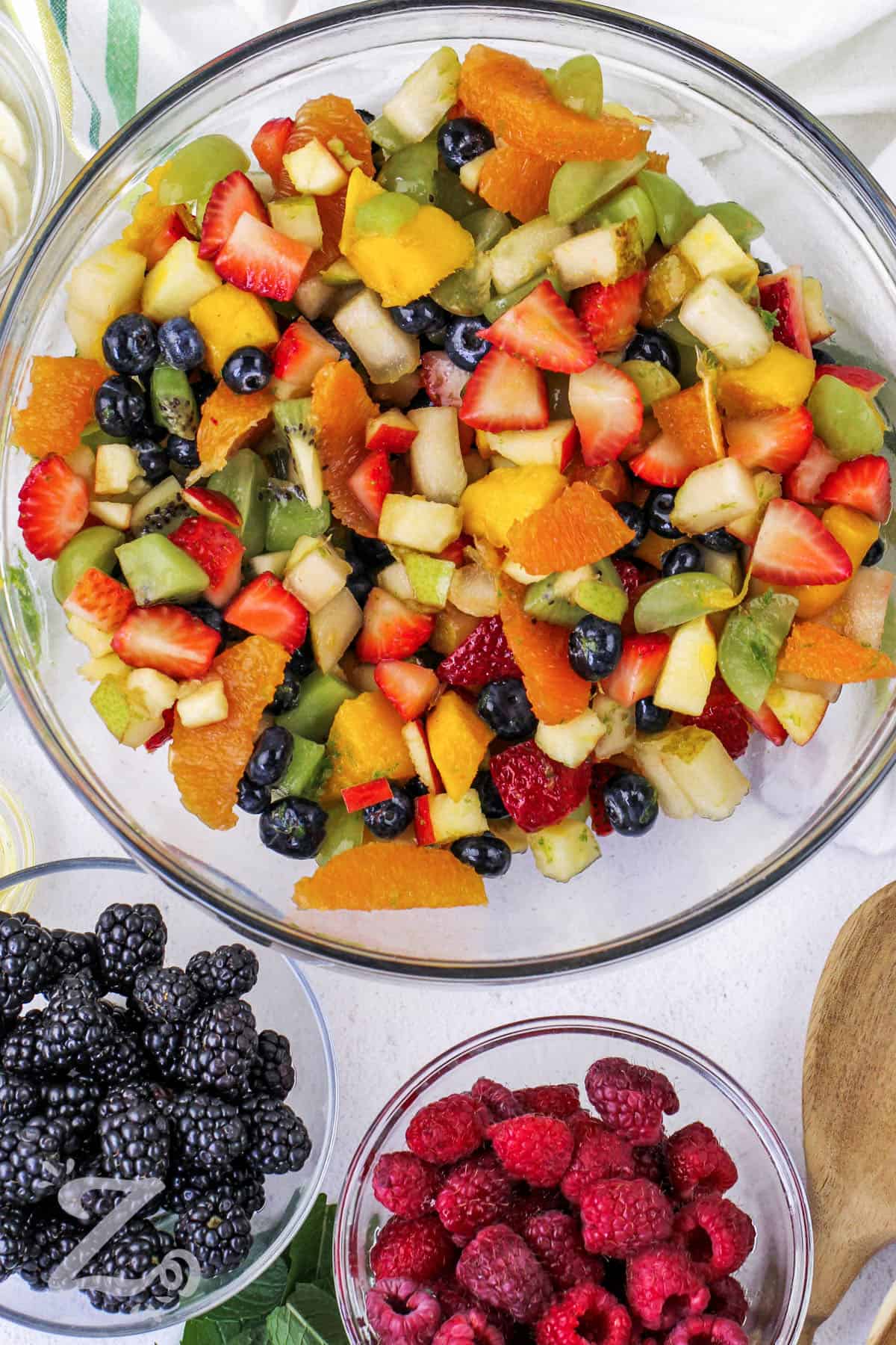 this easy recipe for fruit salad has assorted fruit mixed up in a clear bowl before topping with raspberries and black berries