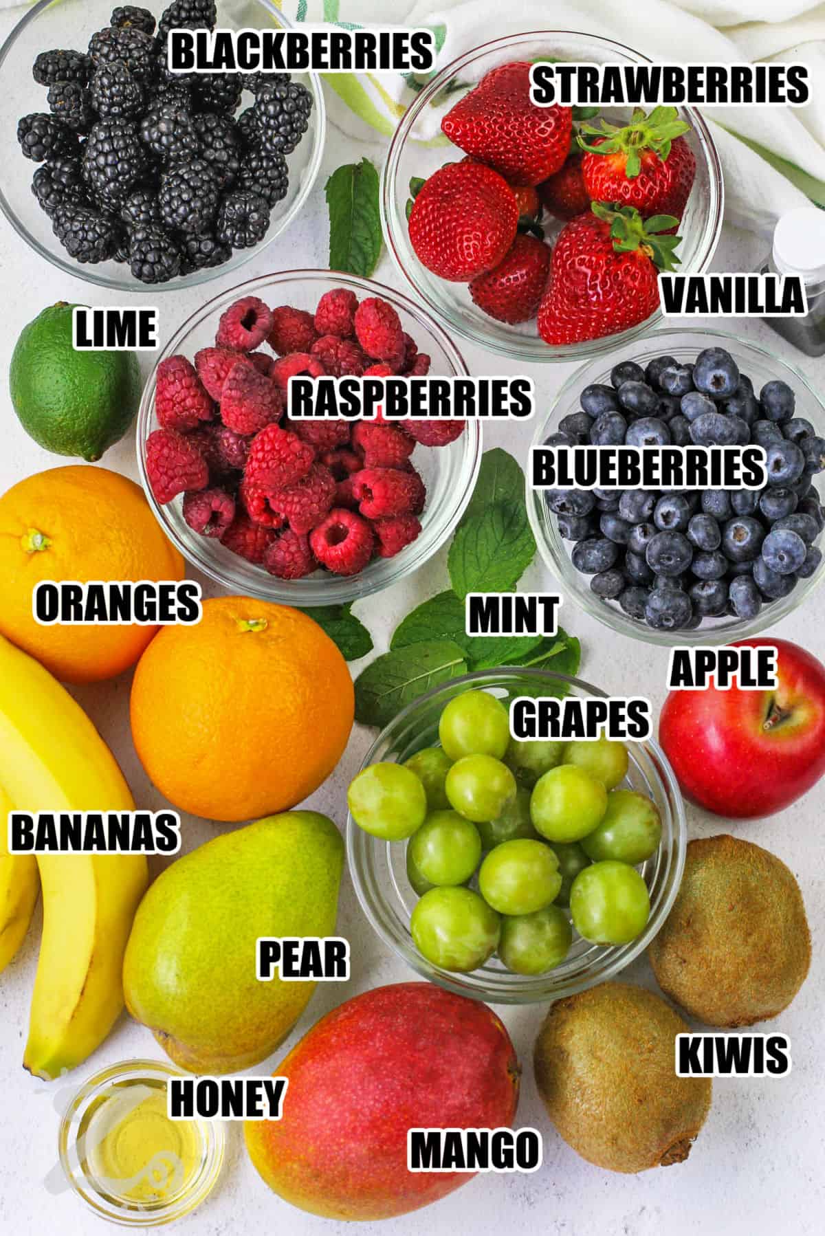 blackberries, strawberries, vanilla, blueberries, apple, grapes, mint, kiwis, mango, honey, pear, bananas, oranges, lime and raspberries assembled to make a recipe for fruit salad.