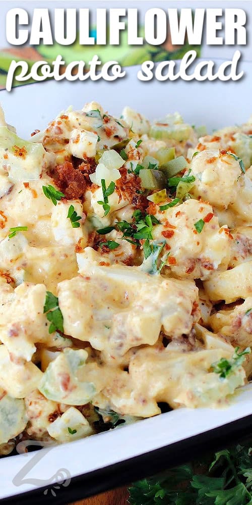 close up of cauliflower potato salad in a white dish, garnished with chopped pickles, bacon bits and chopped parsley with a title