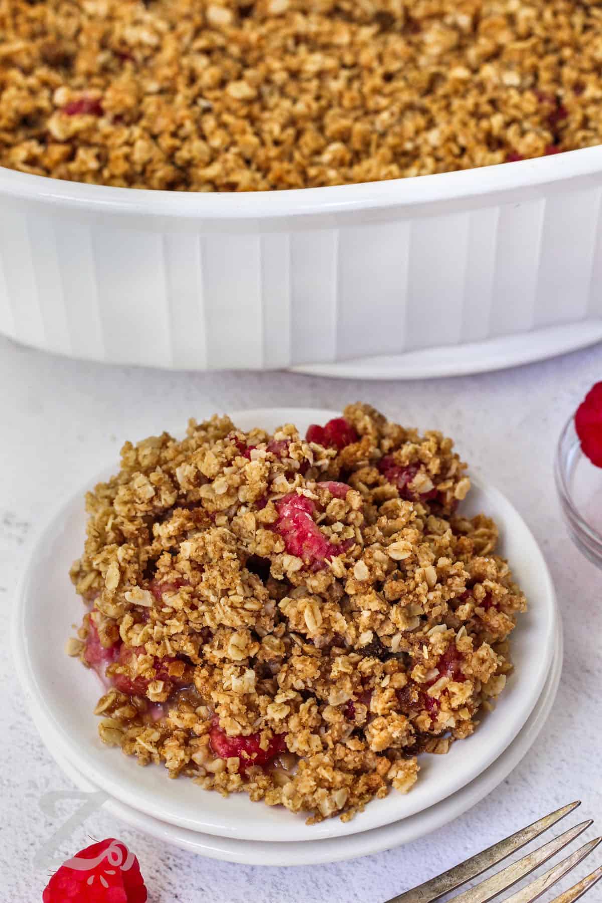 a serving of raspberry crisp on a small white plate, with raspberry crisp in a white baking dish in the background