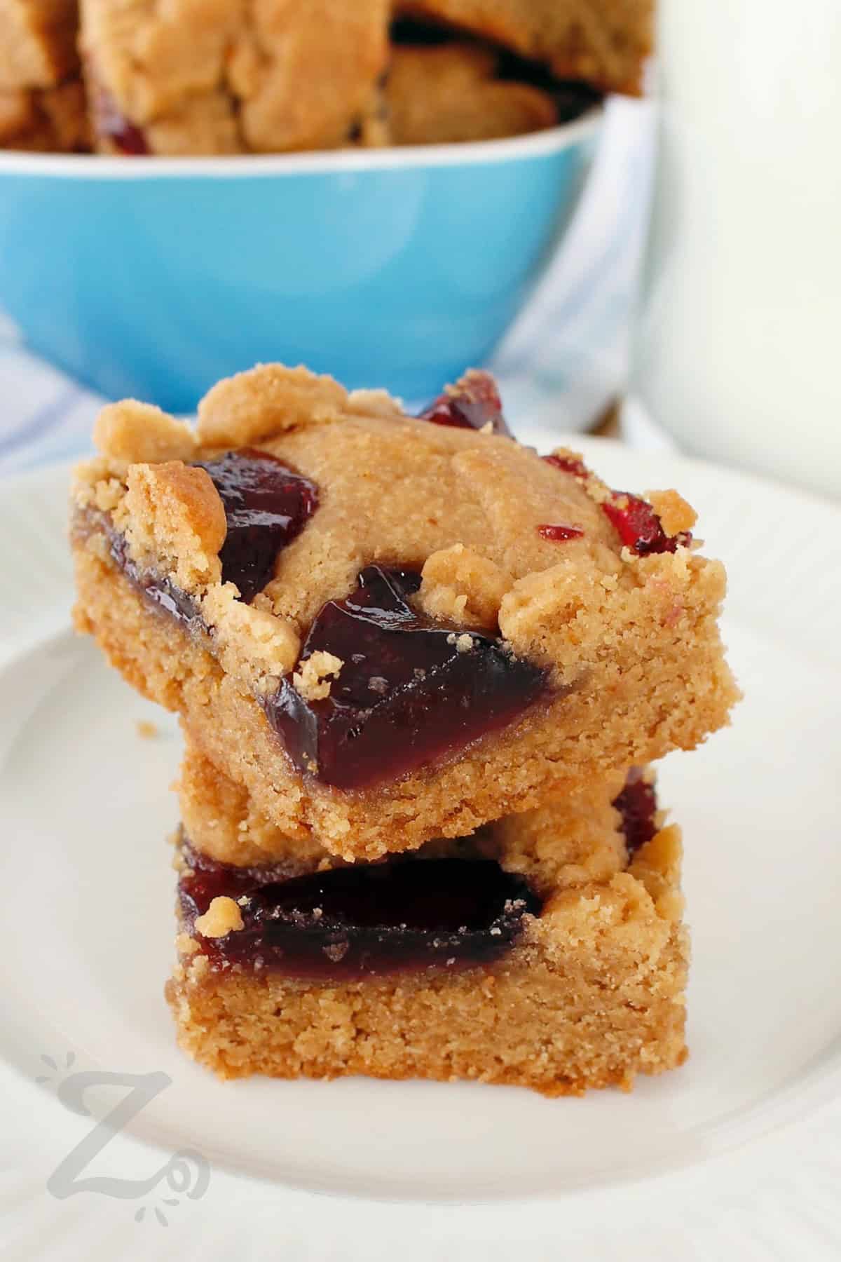 two peanut butter and jelly bars stacked on a white plate, with more bars in a blue bowl in the background