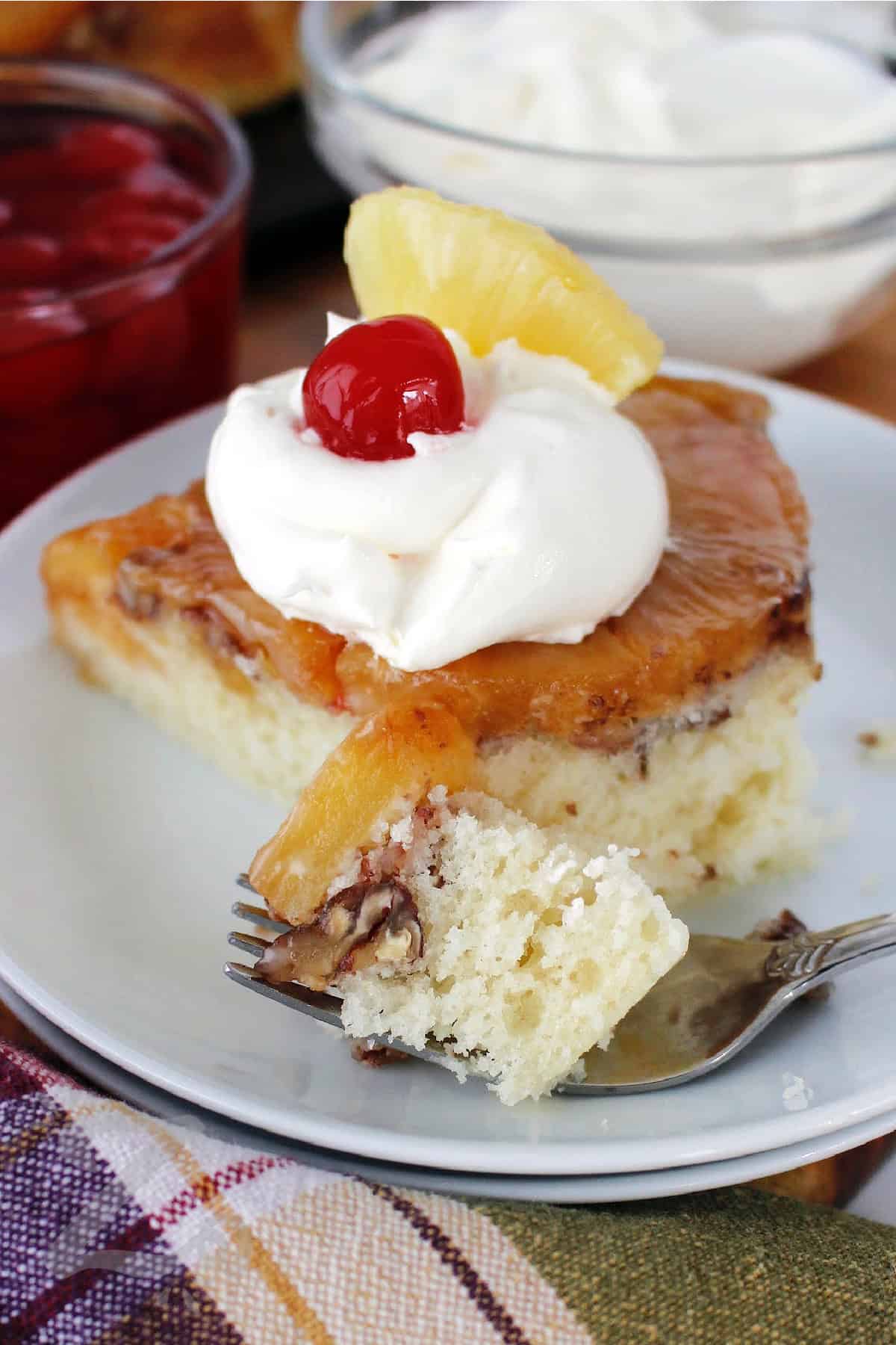 a slice of homemade pineapple upside down cake, garnished with whipped cream, a maraschino cherry, and a chunk of pineapple, on a white plate