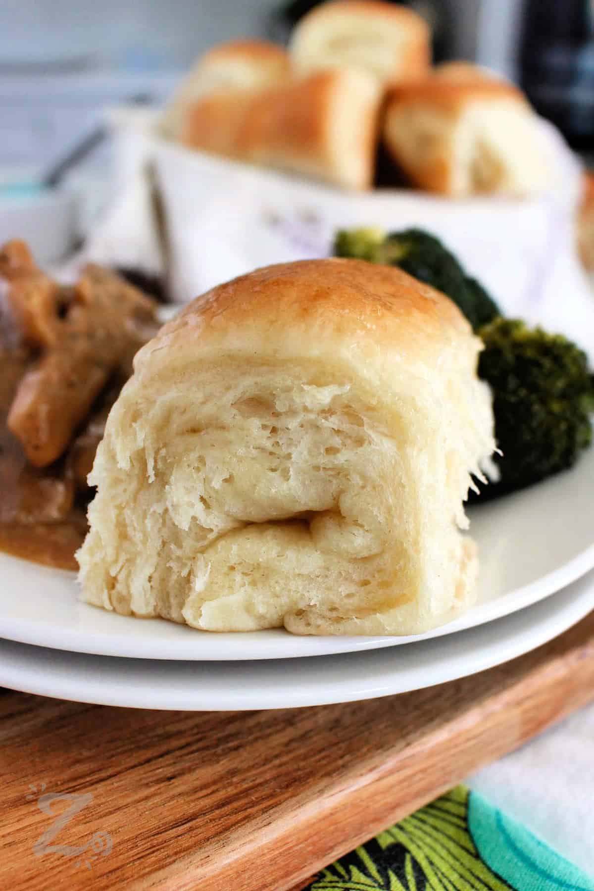 one Parker House Roll served on a white plate with broccoli and pork stew in the background