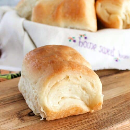 one Parker House Roll sitting on a wooden board, with remaining Parker house rolls in a basket in the background