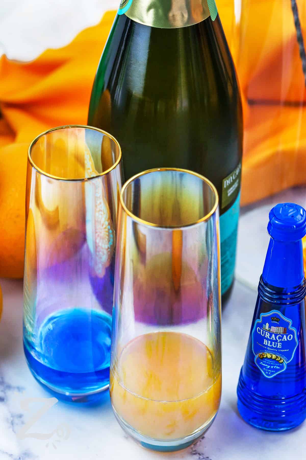 blue curacao in a glass and orange juice in a glass with champagne in the background to make Green Mimosa
