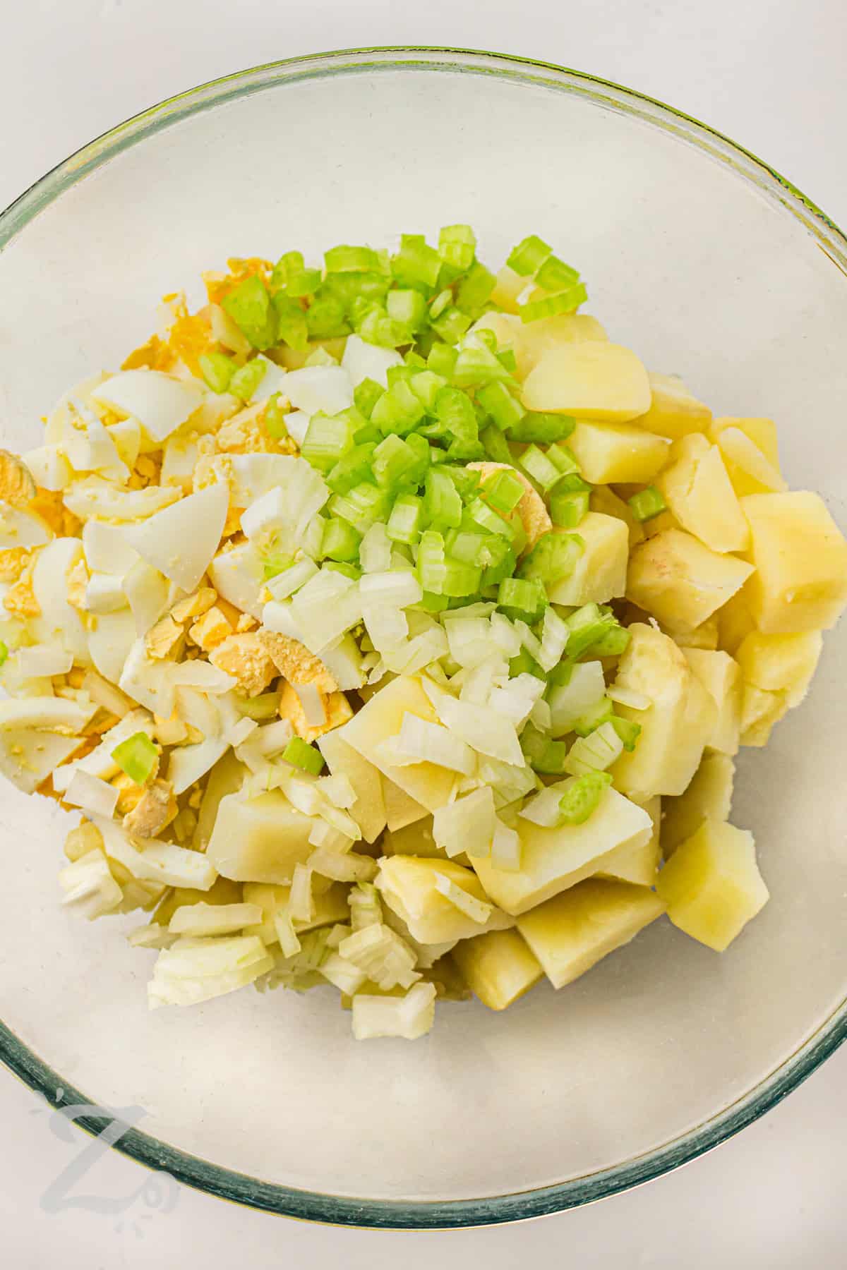 Classic potato salad recipe fans know this creamy side dish belongs on every picnic table. Potatoes, eggs, celery, and onion are mixed with a rich homemade dressing that is smooth, tangy, and full of savory taste. This chilled potato salad is perfect for backyard barbecues, holiday meals, and easy family dinners. Serve it with sandwiches, grilled burgers, or barbecue chicken. #ourzestylife adding ingredients together to make Classic Potato Salad