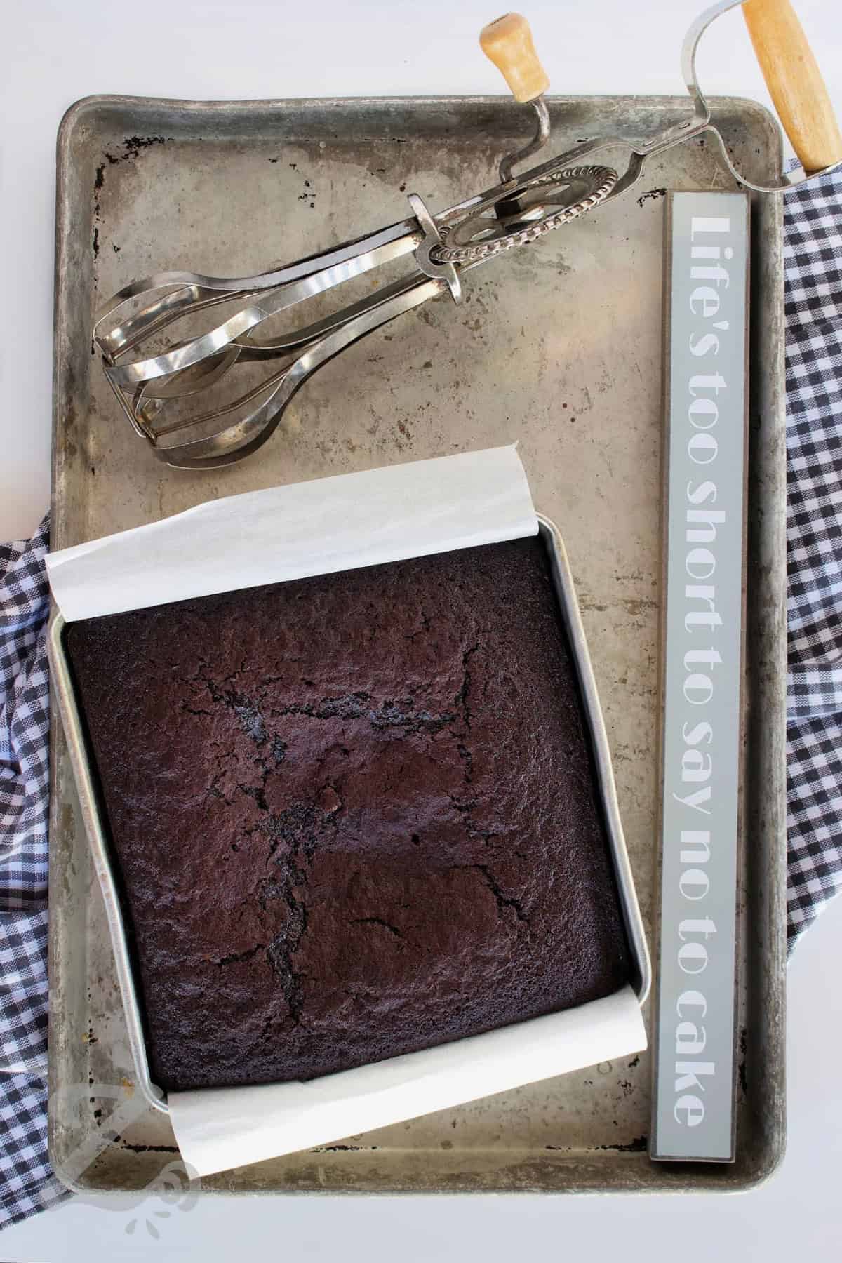 Homemade chocolate cake made simple, rich, and incredibly delicious. With a soft, moist texture and bold cocoa flavor, this recipe is perfect for birthdays, holidays, or anytime you need a quick chocolate fix. It’s the one you’ll come back to again and again so why not try it tonight? #ourzestylife baked homemade chocolate cake in a baking pan on a baking sheet with an old fashioned beater on the side