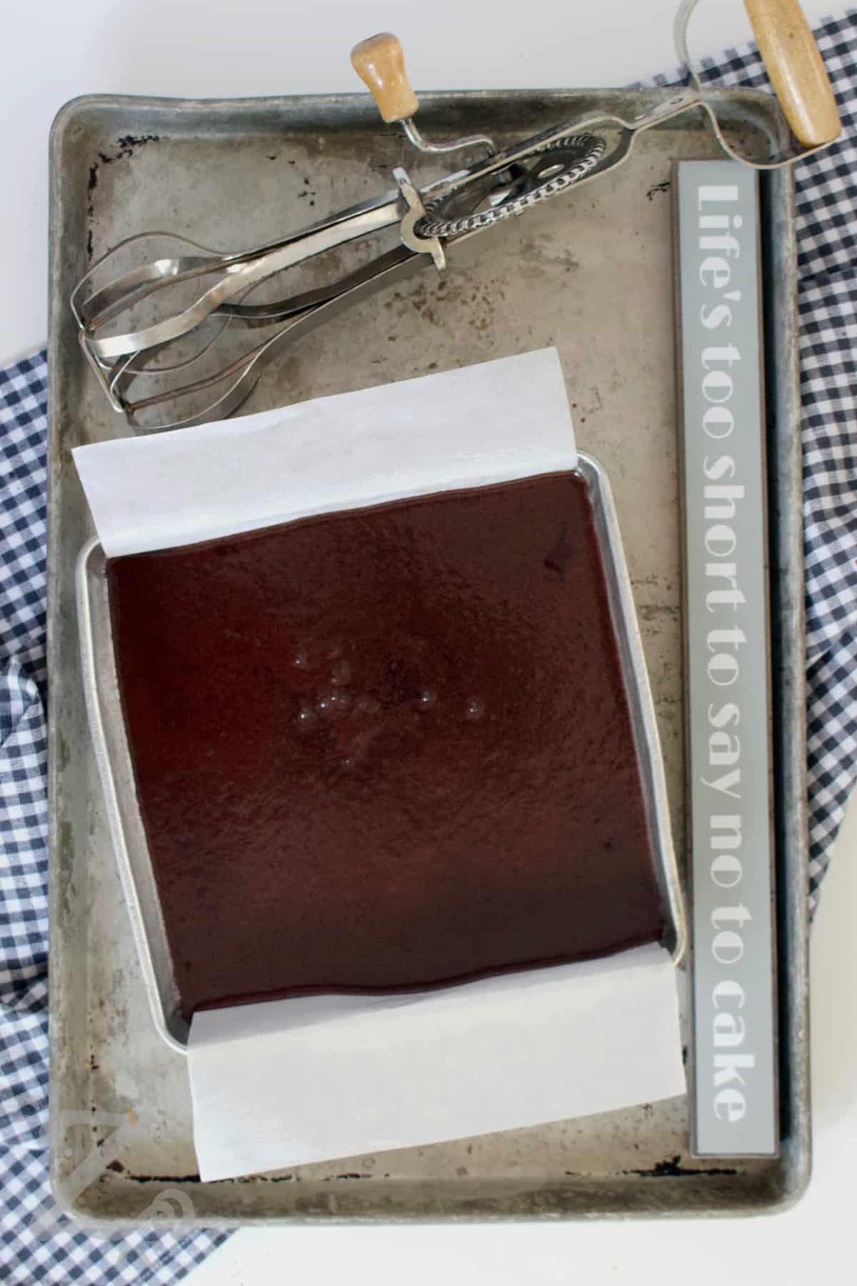 This easy chocolate cake recipe delivers bakery style results with minimal effort. It’s moist, rich, and balanced with deep chocolate flavor in every bite. Ideal to layer, frost with buttercream, or simply enjoy as a snack cake so why not give this recipe a try? #ourzestylife homemade chocolate cake batter in a baking pan on a baking sheet with an old fashioned beater on the side