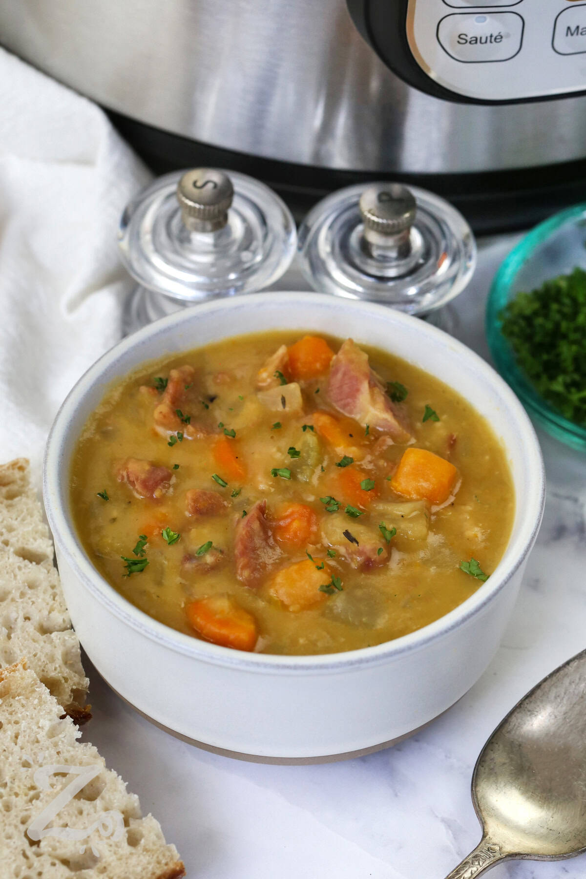 Instant Pot split pea soup is packed with smoky ham, split peas, and tender veggies in every hearty bite. This easy soup recipe creates rich homemade flavor that tastes comforting and satisfying from the very first spoonful. The creamy texture and savory broth make it perfect for chilly evenings when you want a warm family dinner. It is a simple pressure cooker recipe that always delivers big flavor. Cook up this hearty soup recipe tonight. #ourzestylife bowl of Instant Pot Split Pea Soup with Ham with instant pot in the back