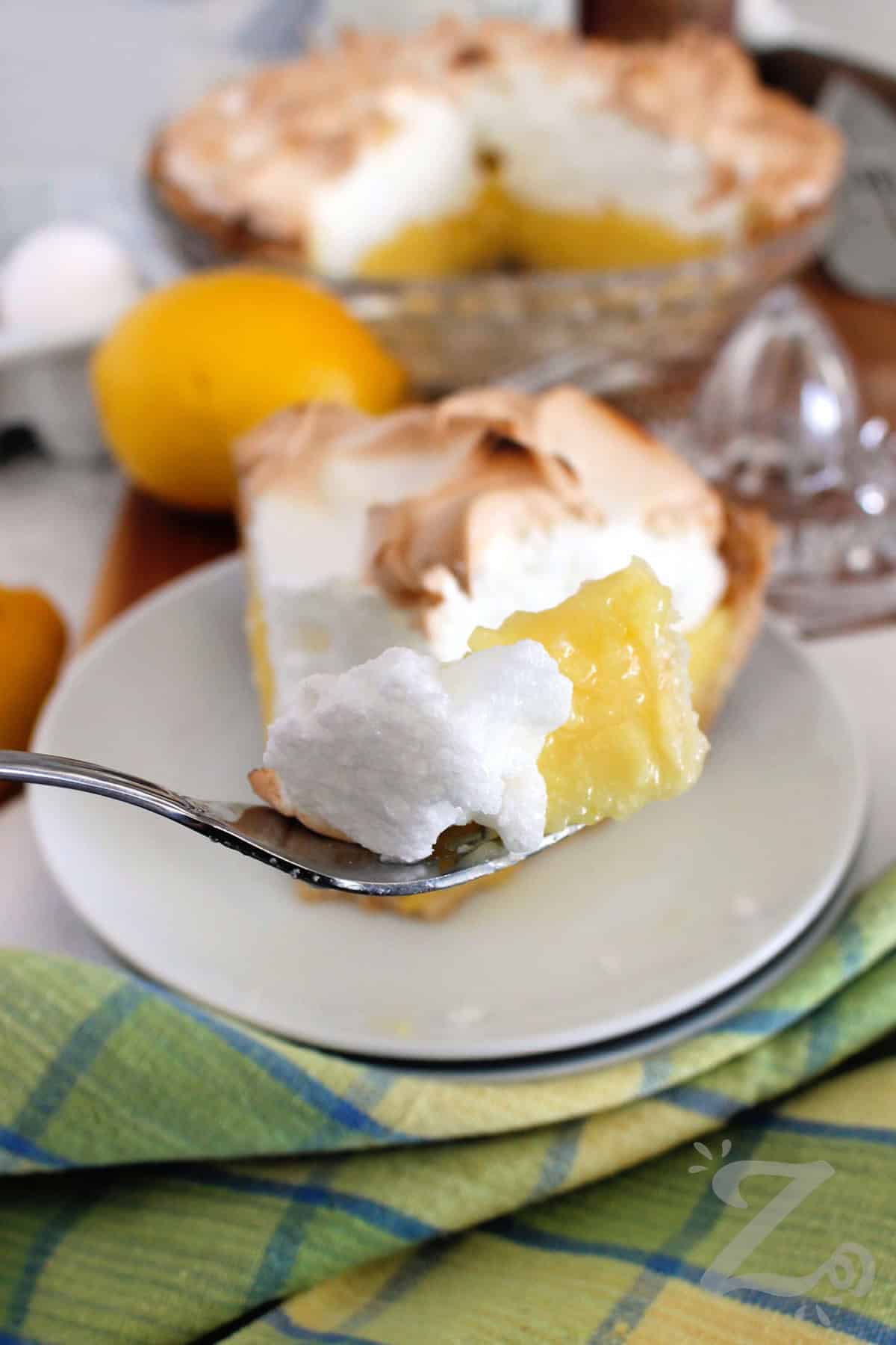 a forkful of lemon meringue pie with an entire pie, eggs, a lemon, and a lemon juicer behind it