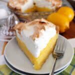 one slice of lemon meringue pie on a white plate, with more pie, lemons, eggs, and lemon juicer in the background