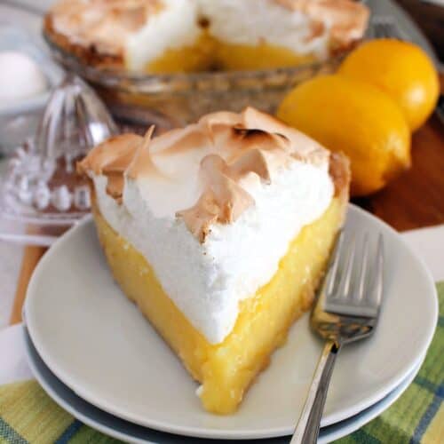 one slice of lemon meringue pie on a white plate, with more pie, lemons, eggs, and lemon juicer in the background
