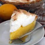 one slice of lemon meringue pie on a white plate, with a whole pie, a lemon, and a lemon juicer behind it