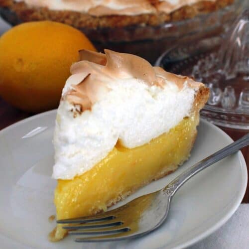 one slice of lemon meringue pie on a white plate, with a whole pie, a lemon, and a lemon juicer behind it
