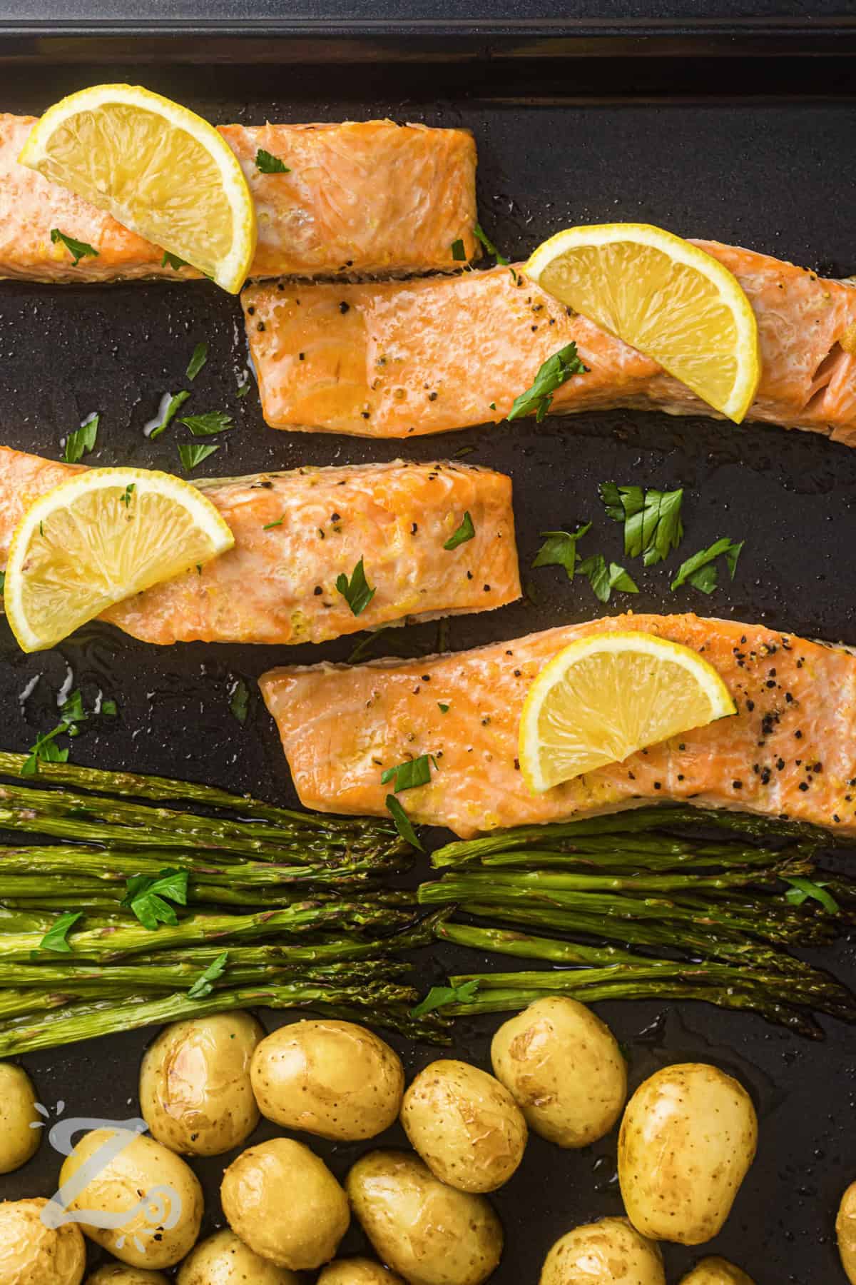 This sheet pan salmon is packed with fresh flavors and simple ingredients that make dinner stress-free. Flaky salmon is coated in a lemon garlic butter mixture and roasted alongside potatoes and asparagus. The result is a colorful meal that is hearty, satisfying, and easy to serve. Keep this one-pan dinner recipe handy for busy nights. #ourzestylife close up of Sheet Pan Salmon with Asparagus and Roasted Potatoes on a sheet pan with lemon slices
