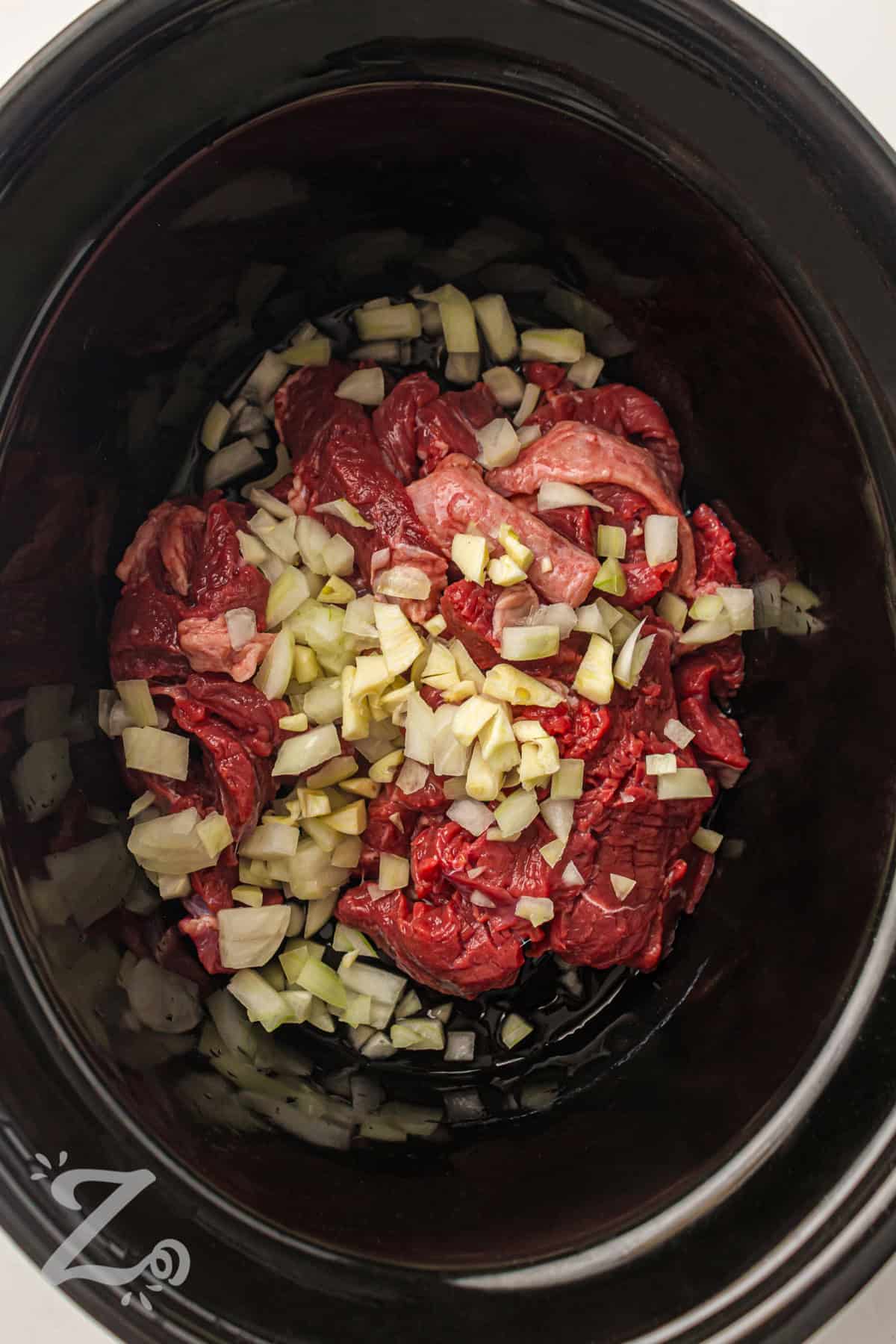 Slow cooker beef and broccoli creates a rich and savory meal that feels like your favorite takeout dish. It combines tender sliced beef with a garlic soy sauce that soaks into every bite. Brown sugar adds a touch of sweetness that balances the flavors perfectly. Serve it over rice or noodles for an easy, complete dinner. Try this recipe tonight for a simple and satisfying meal. #ourzestylife adding garlic and onions to beef to make Slow Cooker Beef and Broccoli