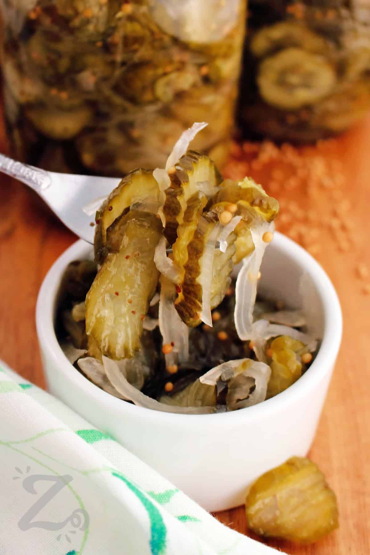 Try this Bread and Butter Pickles recipe at home and enjoy a sweet and tangy condiment on your favorite dishes. The recipe is easy enough for first-time canners or someone who just wants quick pickles stored fresh in the refrigerator. It’s a recipe that creates a delicious way to preserve fresh cucumbers and enjoy them later. #ourzestylife bread and butter pickles recipe on a fork