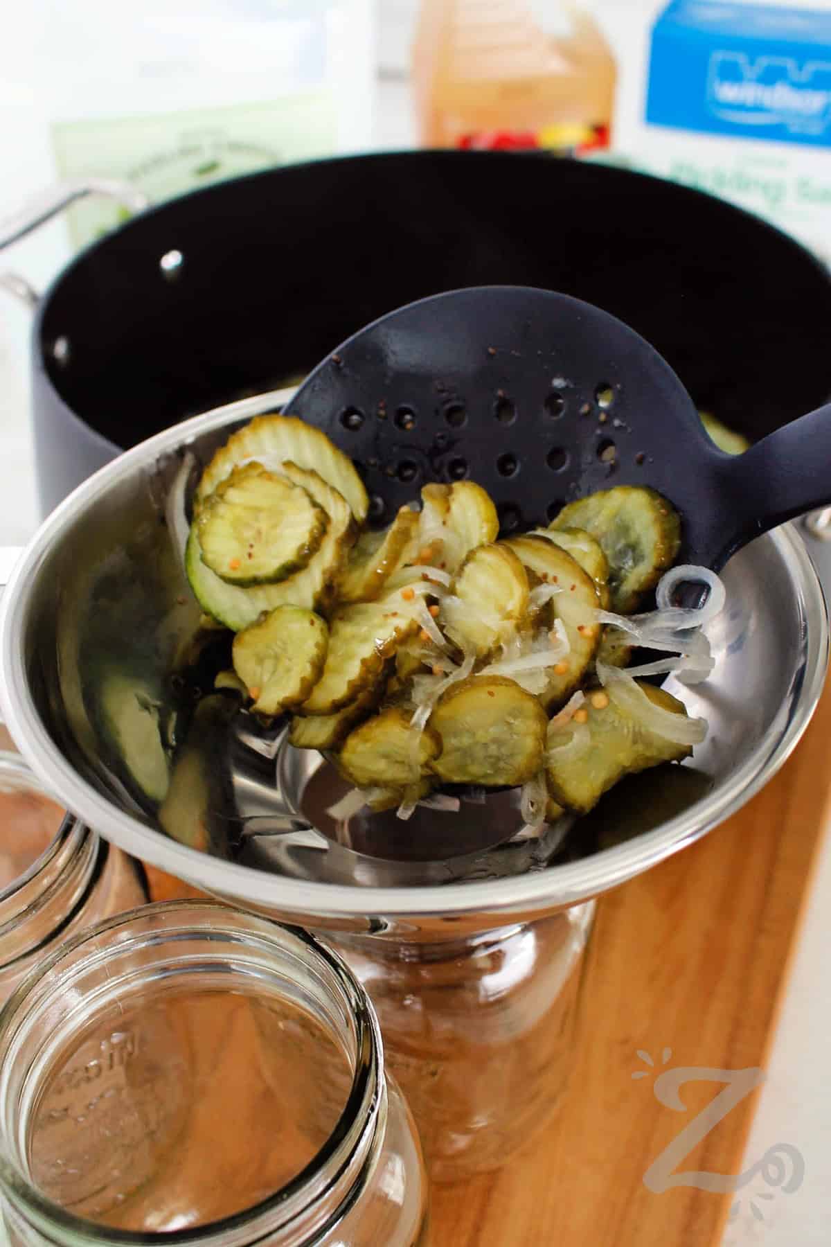 This Bread and Butter Pickles recipe is one you need to try. The pickles are sweet, crunchy, and bursting with homemade flavor, and the recipe is easy enough for first-time canners or someone who just wants quick pickles stored in the refrigerator. It’s a recipe that creates a delicious way to preserve fresh cucumbers and enjoy them later. #ourzestylife process to make bread and butter pickles recipe where cucumbers and onions are being ladled into a mason jar through a wide mouth funnel