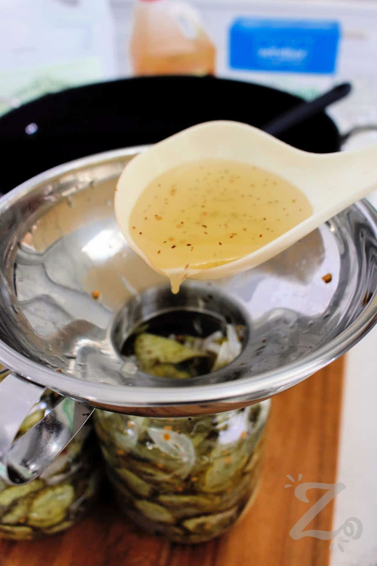 Make a batch of homemade Bread and Butter Pickles and enjoy garden-fresh flavor all year long. This easy recipe creates crisp, sweet pickles with just the right balance of vinegar, sugar, and spices. Or modify it and create refrigerator pickles you can enjoy in 3 months time. Either way, this is a must try recipe. #ourzestylife process to make bread and butter pickles recipe where the brine is being poured into mason jars using a wide mouth funnel