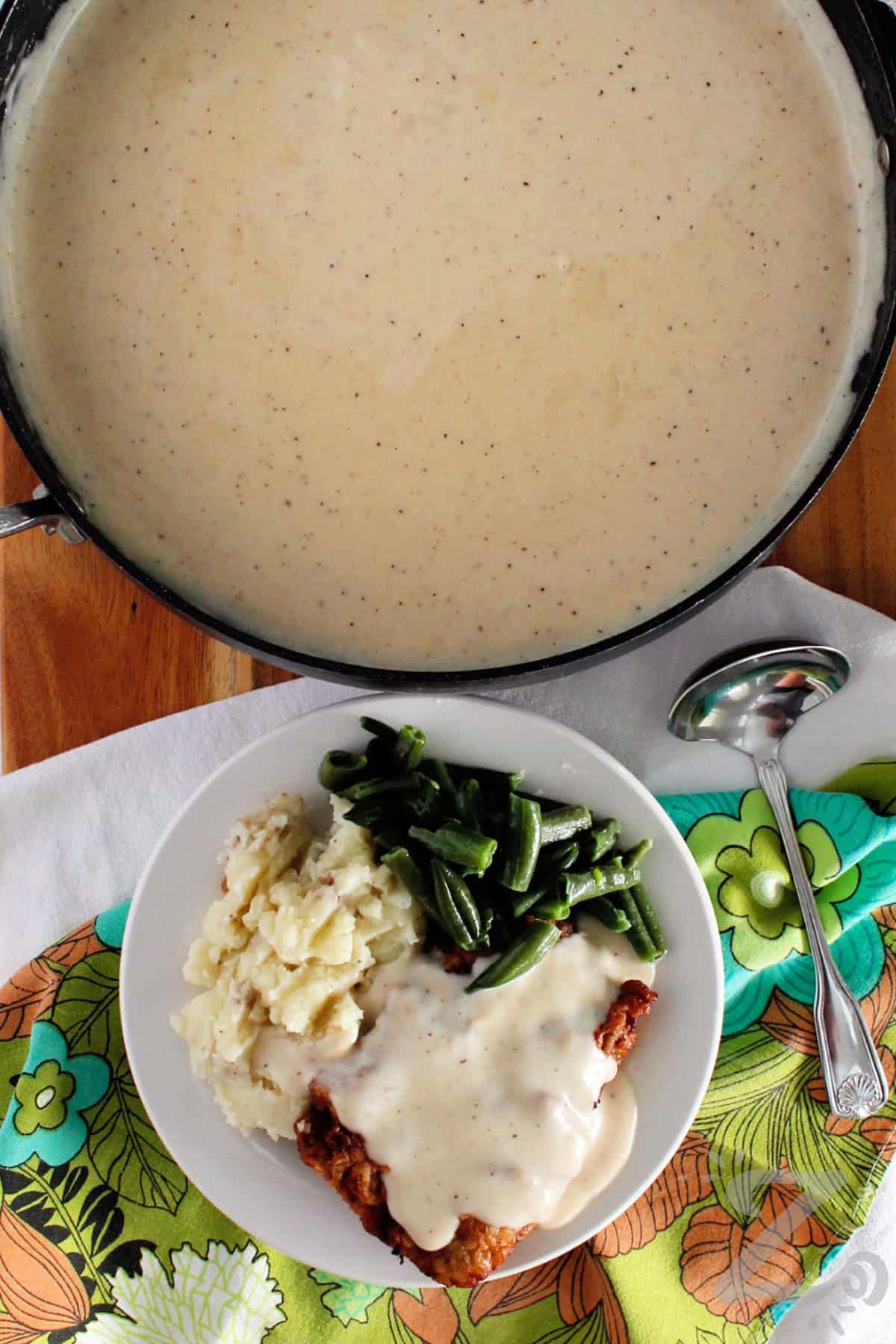 chicken fried steak with country gravy on a white plate with mashed potatoes, green beans, and gravy in a saucepan on the side