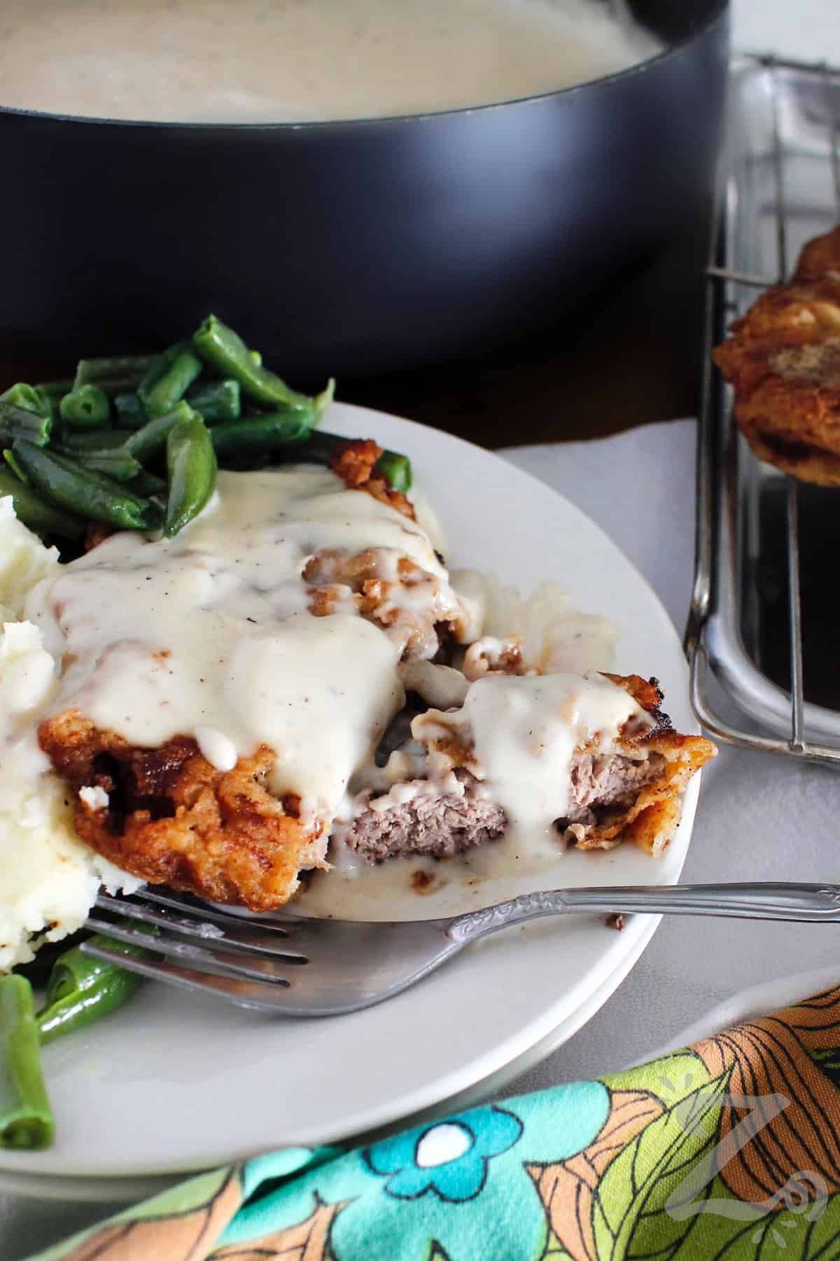 chicken fried steak with country gravy, with a small bite cut out, on a white plate with mashed potatoes and green beans