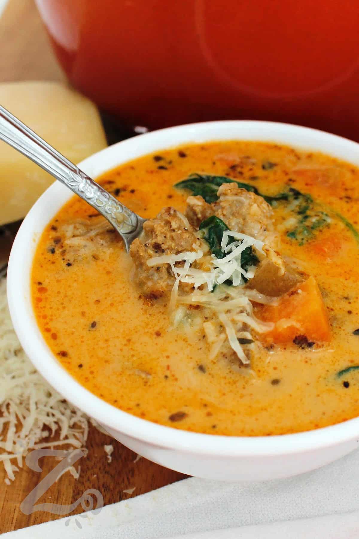 This Sausage Potato Soup is a cozy one-pot meal with Italian sausage, tender potatoes, carrots, celery, spinach, in a creamy broth. A touch of red pepper adds just enough heat to balance the rich, savory flavors. Sausage and potato soup is hearty enough for a chilly weeknight meal so why not make a pot tonight? #ourzestylife sausage potato soup in a white bowl with shredded parmesan garnish and a spoon.