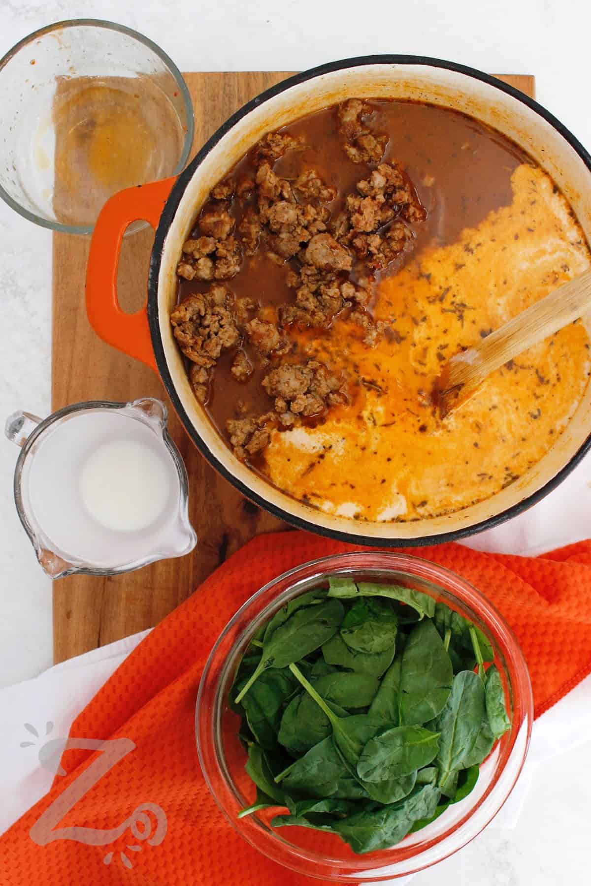 Sausage Potato Soup turns simple ingredients into a satisfying comfort food meal. Rich Italian sausage, tender potatoes, sweet carrots, savory celery, and fresh spinach come together in a creamy broth with a little heat. This sausage and potato soup is perfect for a cozy night at home, so why not give it a try! #ourzestylife sausage potato soup in a pot when cream and sausage has been added, with spinach on the side.
