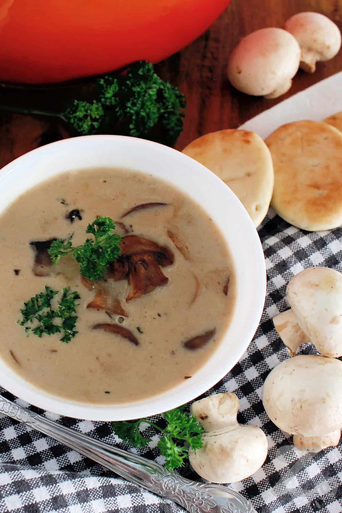 Homemade cream of mushroom soup in a white bowl garnished with fresh parsley, mushrooms, pitas, and a spoon on the side.